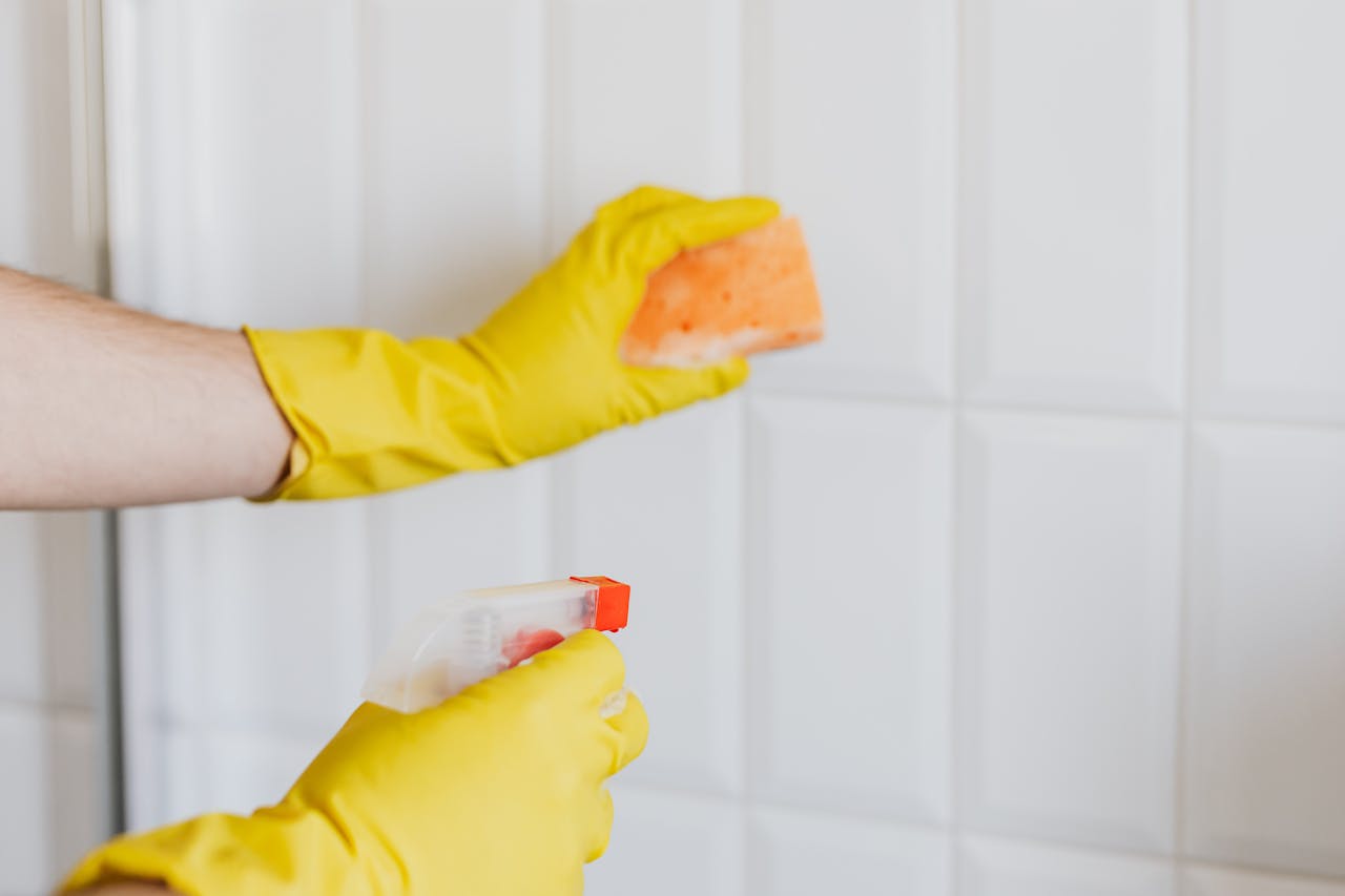 hero-img A person wearing yellow gloves cleans bathroom tiles with a sponge and spray bottle.